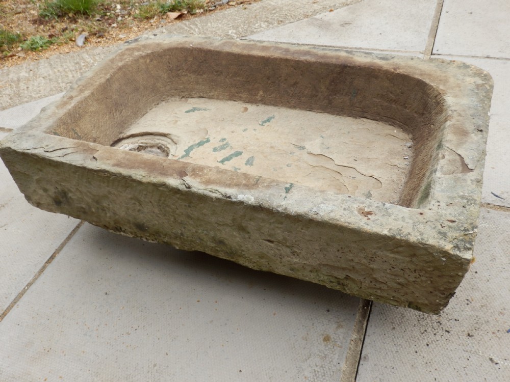 A rectangular, hand-carved antique stone sink displaying a textured exterior, weathered patina, and a corner drainage hole.