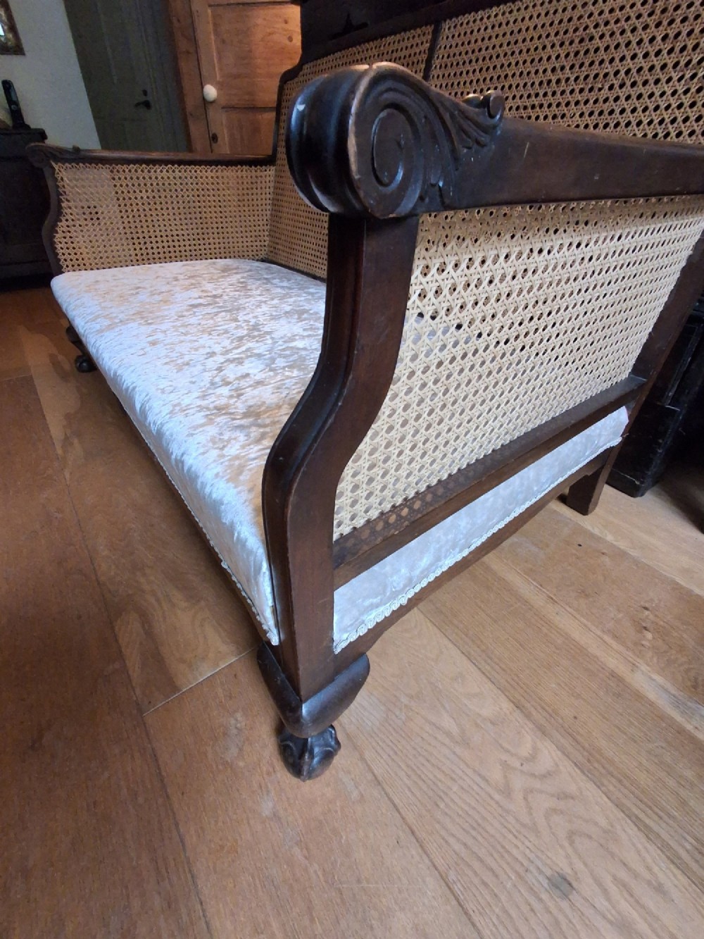 A French style two-seater sofa featuring a dark carved wooden frame with woven cane side panels and light cream velvet seat and back cushions. View III