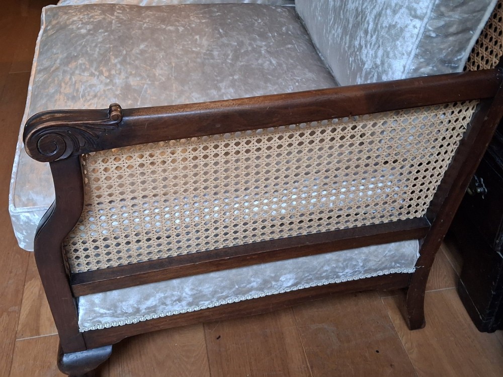 A French style two-seater sofa featuring a dark carved wooden frame with woven cane side panels and light cream velvet seat and back cushions. View IV