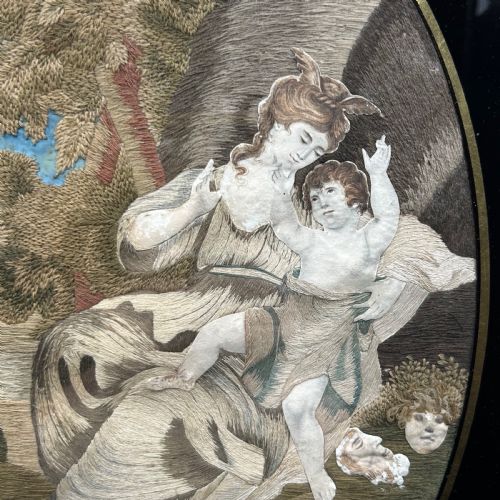 fig 2. An oval Georgian silk embroidery depicting a seated woman and child within a landscape, featuring painted paper faces and set in a black frame.