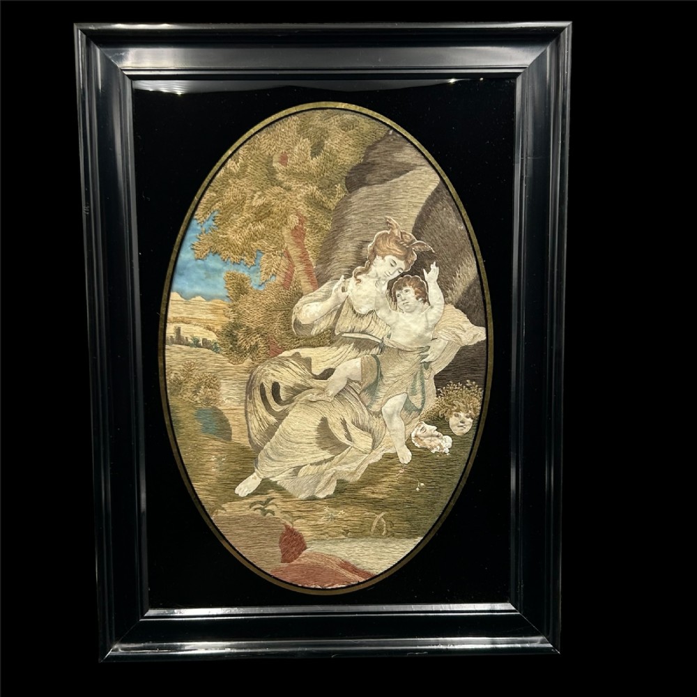 An oval Georgian silk embroidery depicting a seated woman and child within a landscape, featuring painted paper faces and set in a black frame.
