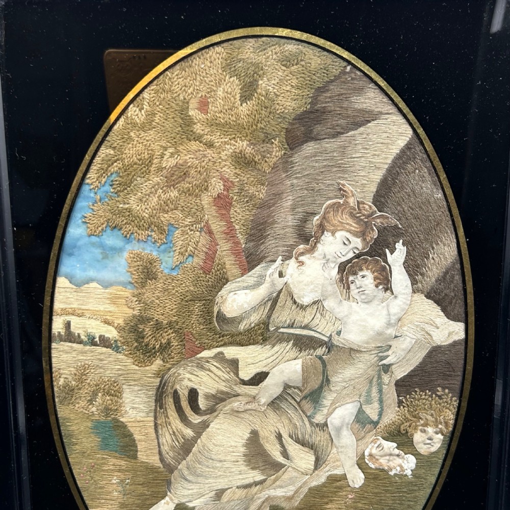 An oval Georgian silk embroidery depicting a seated woman and child within a landscape, featuring painted paper faces and set in a black frame. View II