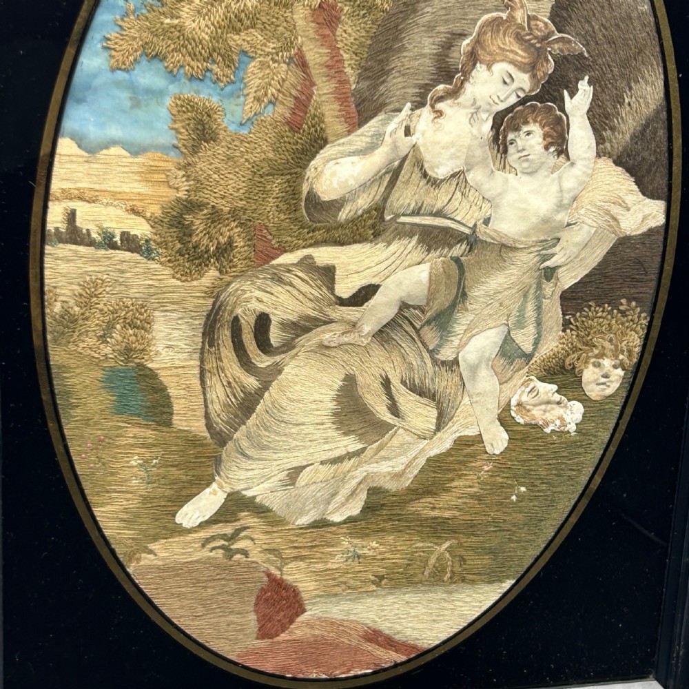 An oval Georgian silk embroidery depicting a seated woman and child within a landscape, featuring painted paper faces and set in a black frame. View V