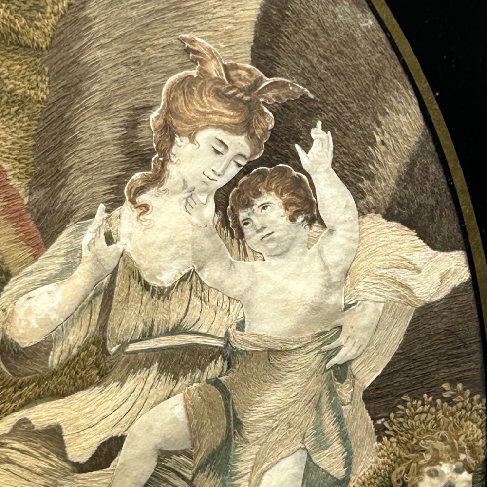 An oval Georgian silk embroidery depicting a seated woman and child within a landscape, featuring painted paper faces and set in a black frame. View VI