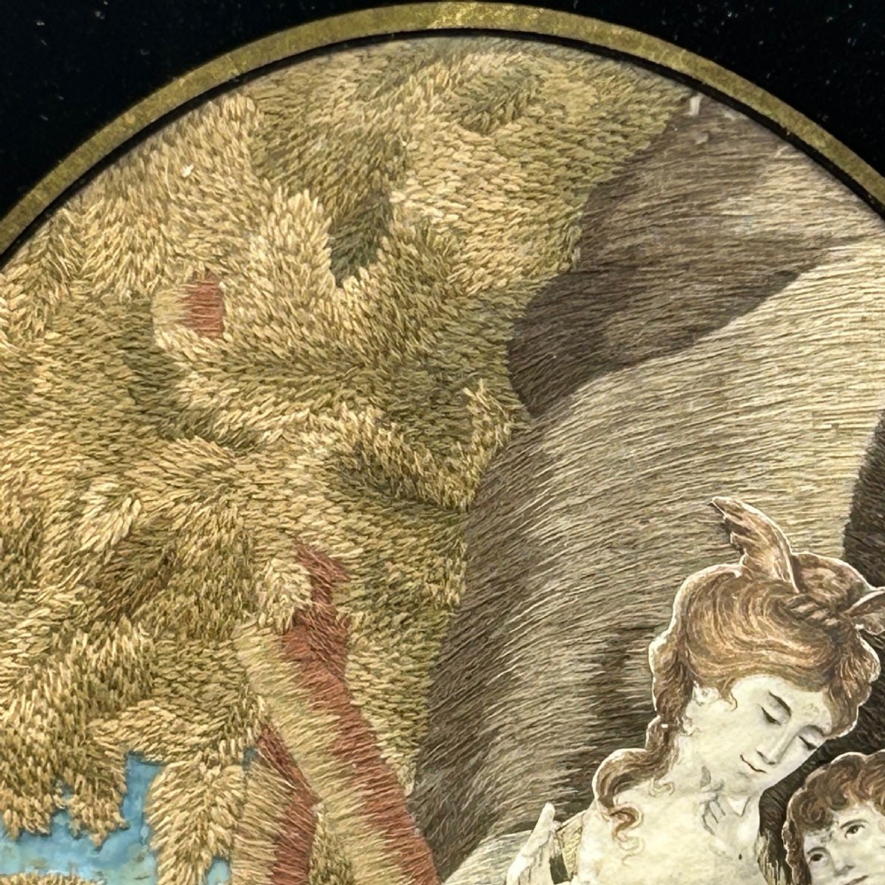 An oval Georgian silk embroidery depicting a seated woman and child within a landscape, featuring painted paper faces and set in a black frame. View VII