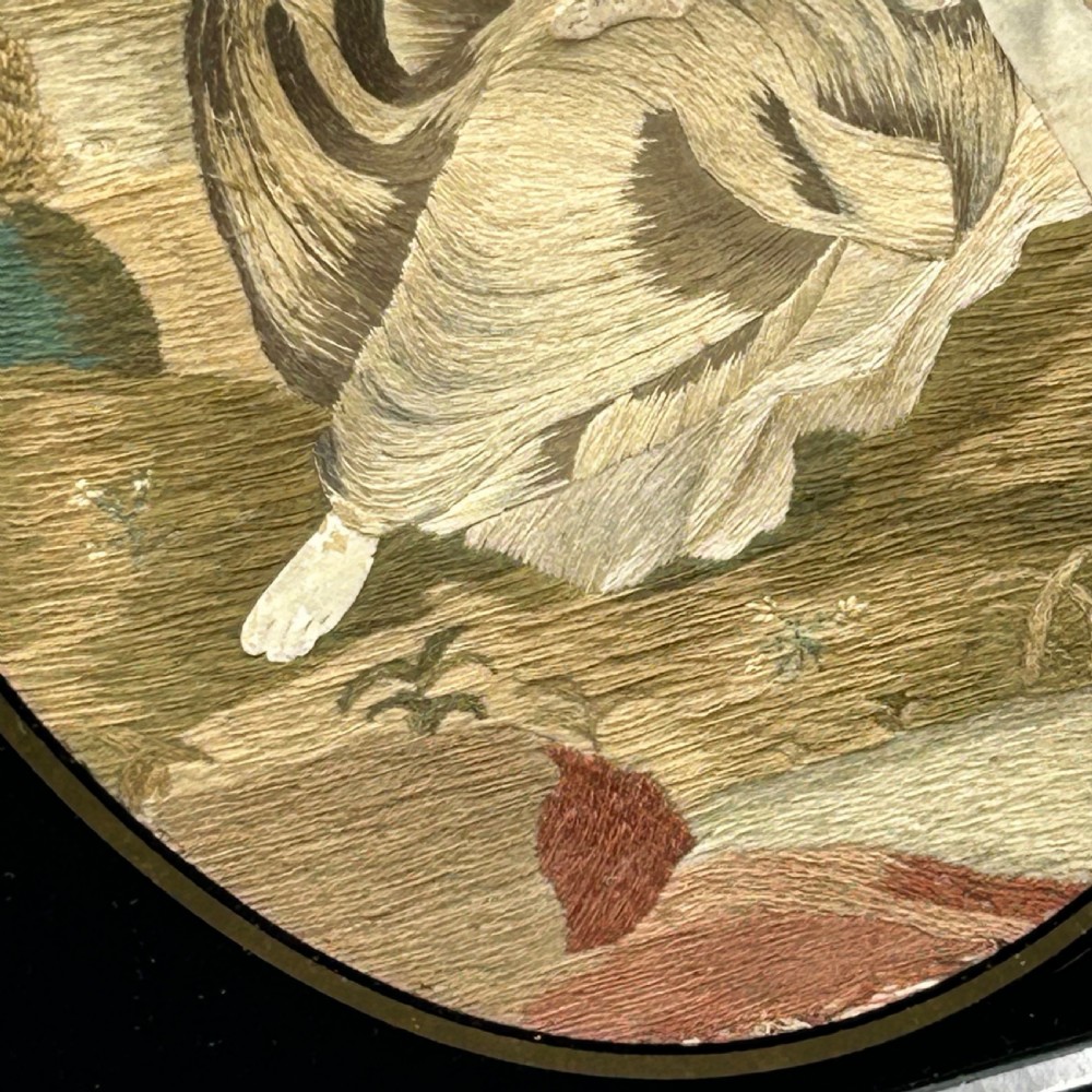 An oval Georgian silk embroidery depicting a seated woman and child within a landscape, featuring painted paper faces and set in a black frame. View IX