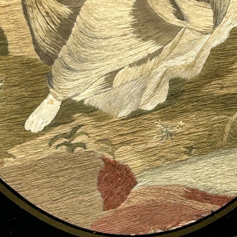 An oval Georgian silk embroidery depicting a seated woman and child within a landscape, featuring painted paper faces and set in a black frame. View X