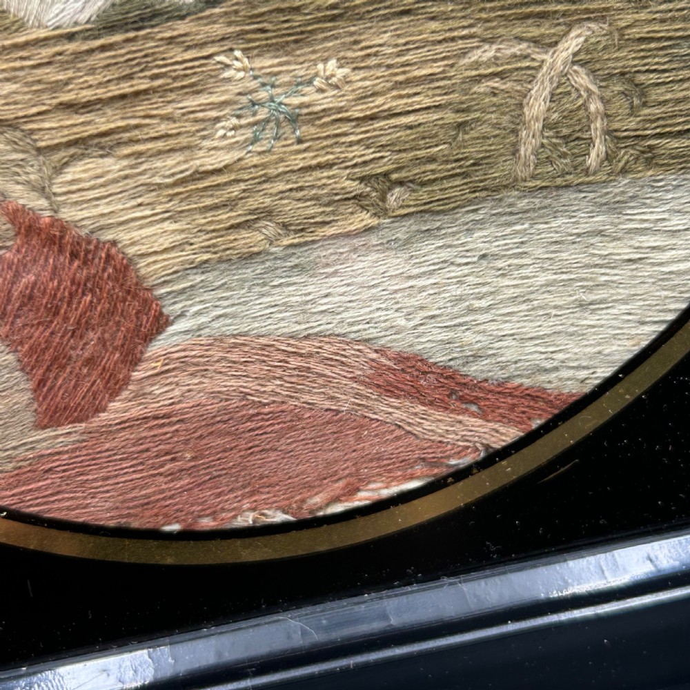 An oval Georgian silk embroidery depicting a seated woman and child within a landscape, featuring painted paper faces and set in a black frame. View XV