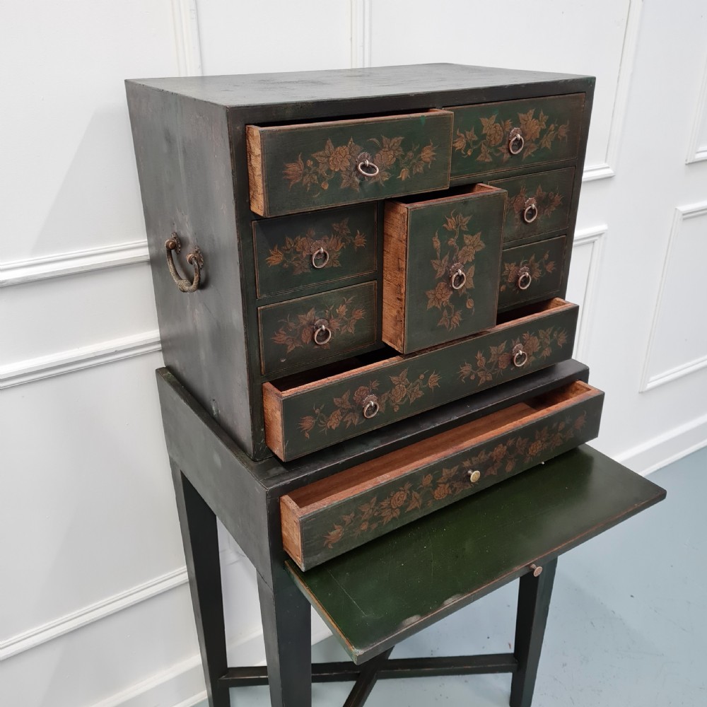 A dark green late Victorian collectors cabinet on a tall stand, featuring hand-painted floral motifs, multiple drawers, and brass ring handles. View III