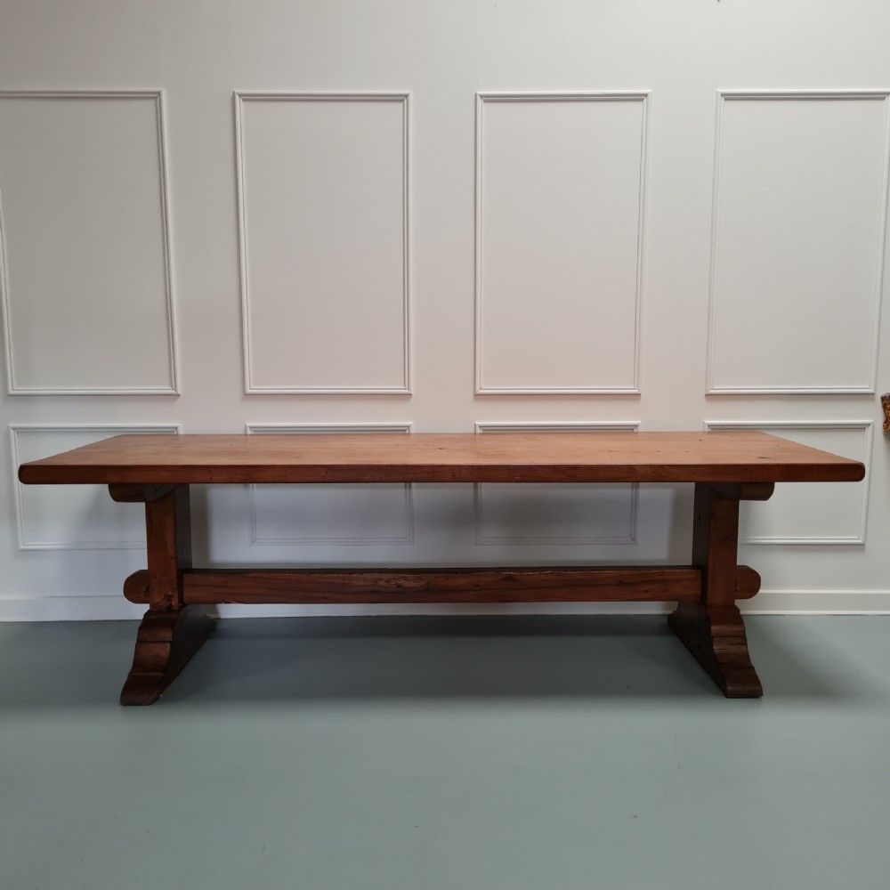 A large, solid warm brown oak monastery dining table featuring a thick rectangular top supported by robust end trestles and a low central stretcher rail.