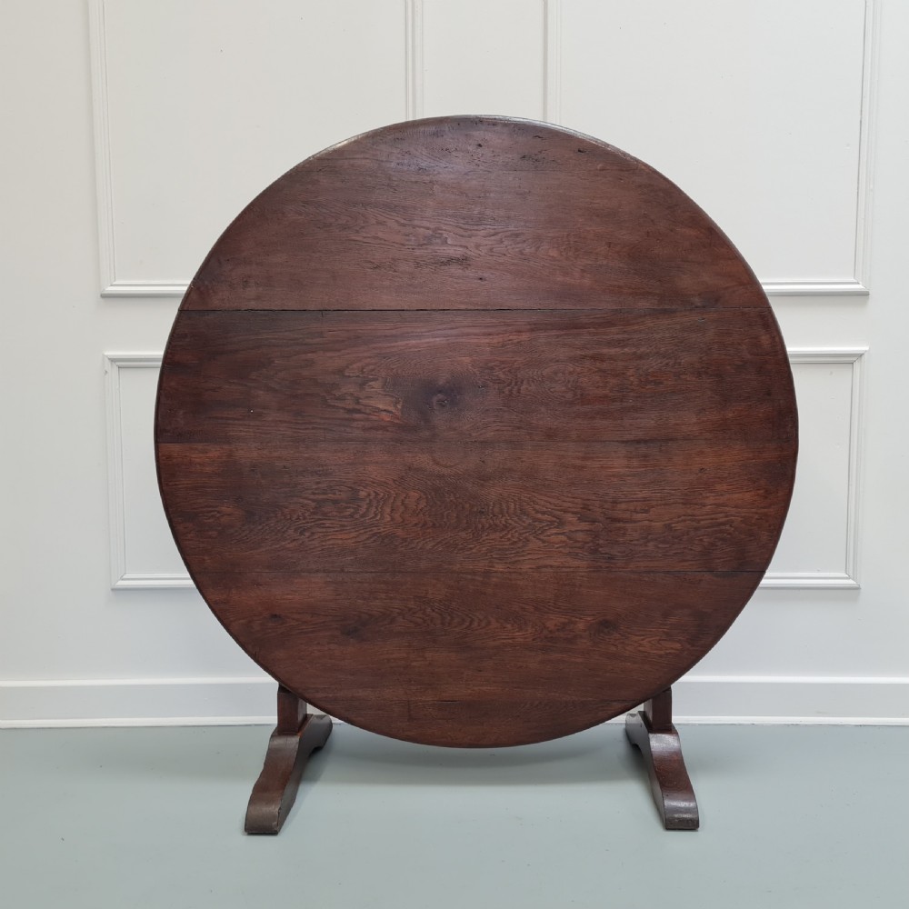 A circular dark oak Vendange table shown with its top tilted vertically on a low wooden trestle base.