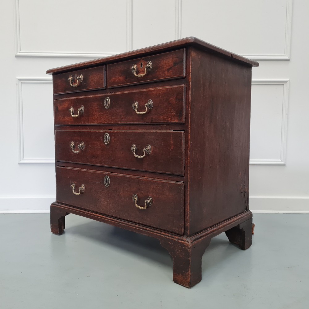 A dark oak Georgian chest of drawers featuring two short drawers over three long drawers with brass swan-neck handles and bracket feet. View V