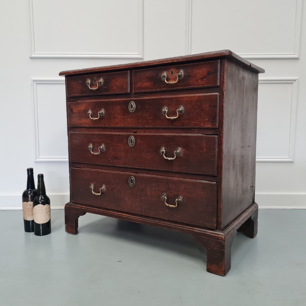A dark oak Georgian chest of drawers featuring two short drawers over three long drawers with brass swan-neck handles and bracket feet. View X