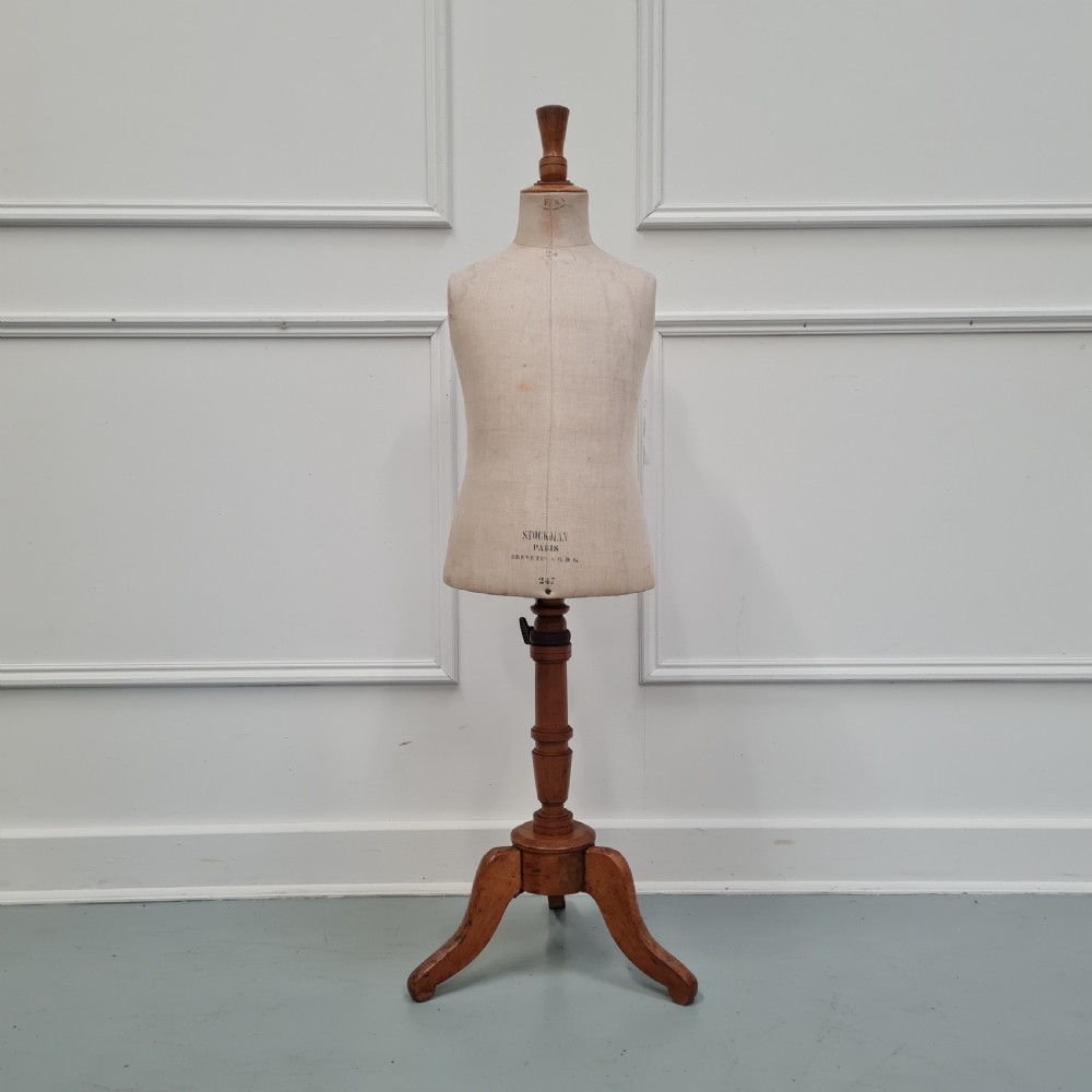 An antique French child-sized tailor mannequin by Stockman featuring a beige linen torso mounted on a turned wooden tripod stand.