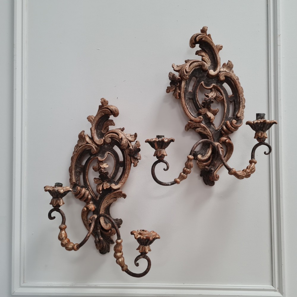 A pair of carved wood wall sconces featuring ornate scrollwork, aged silver leaf finish, and twin candle arms.