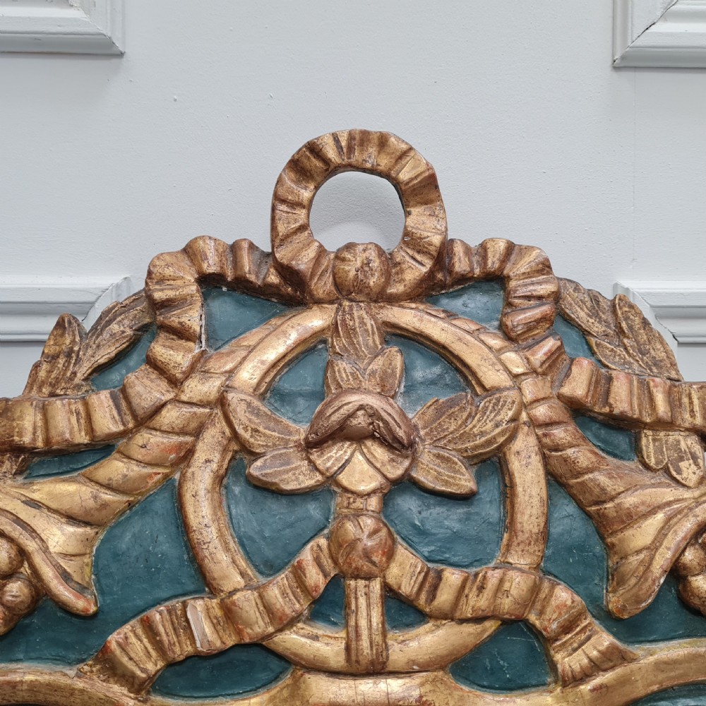 An 18th-century French gilded wooden mirror featuring a carved ornate crest with bow and garland motifs set against a blue background, enclosing a foxed rectangular mirror plate. View IV