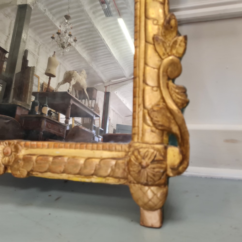 An 18th-century French gilded wooden mirror featuring a carved ornate crest with bow and garland motifs set against a blue background, enclosing a foxed rectangular mirror plate. View V
