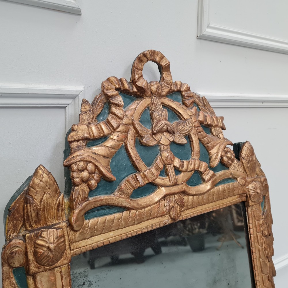 An 18th-century French gilded wooden mirror featuring a carved ornate crest with bow and garland motifs set against a blue background, enclosing a foxed rectangular mirror plate. View VI