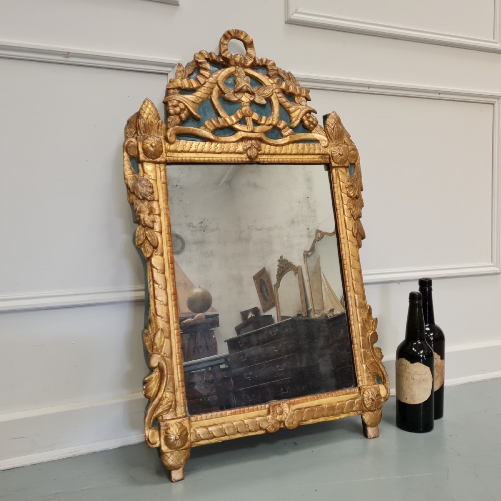 An 18th-century French gilded wooden mirror featuring a carved ornate crest with bow and garland motifs set against a blue background, enclosing a foxed rectangular mirror plate. View VII