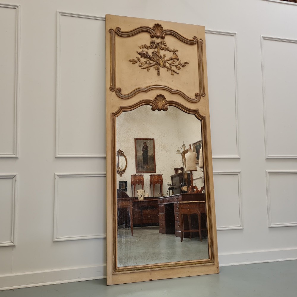 A tall French trumeau mirror featuring a cream painted frame with gilt carved moulding, an upper relief panel displaying a trophy motif, and rectangular glass below.