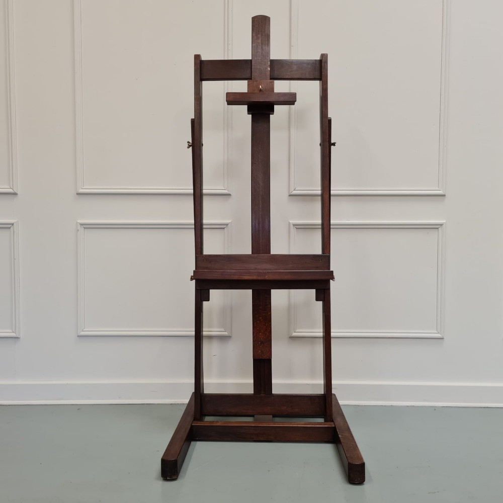 A dark brown French oak H-frame studio easel featuring an adjustable canvas shelf and metal side fixtures.