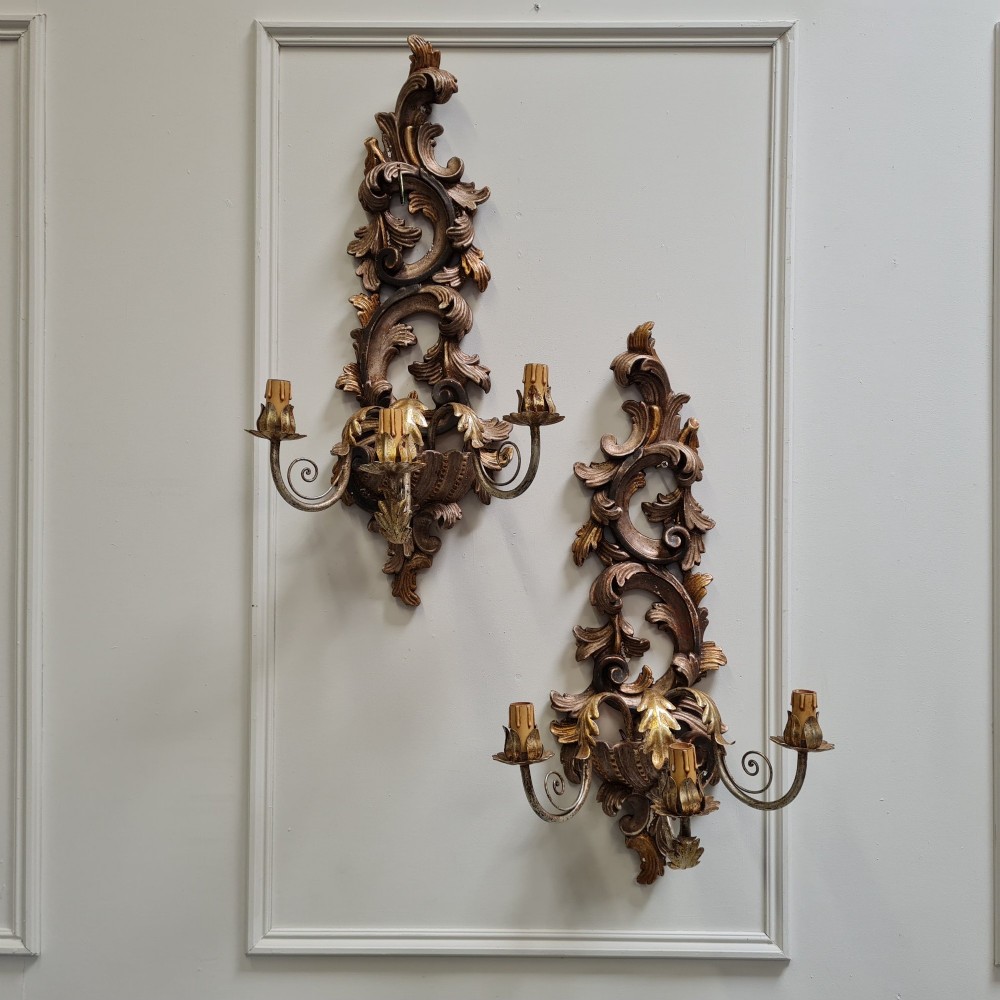 A pair of carved wooden Italian wall sconces featuring ornate acanthus leaf scrolls, gilded highlights, and three metal candle-style light branches.