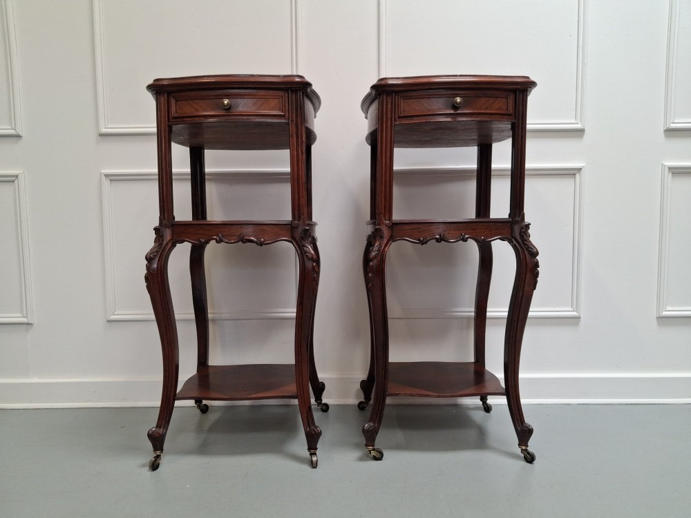 A matching pair of tall dark wood end tables featuring top drawers, lower open shelves, carved cabriole legs, and brass castors.