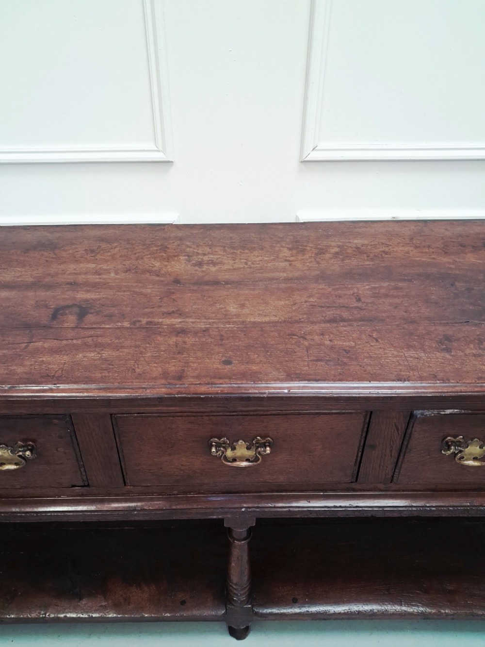 An 18th-century dark oak Georgian dresser base featuring three drawers with brass handles, turned front legs, and a low pot board shelf. View II