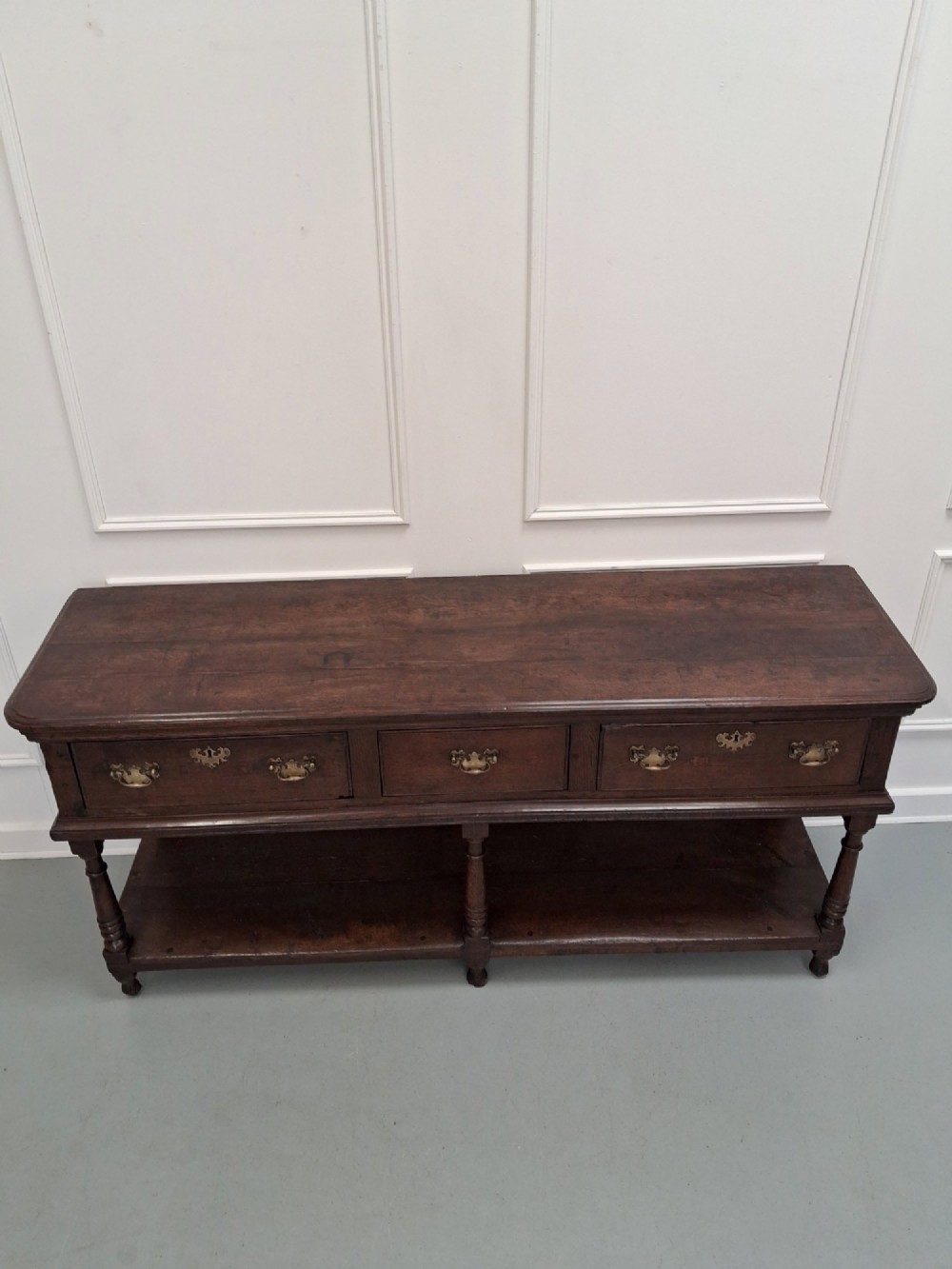 An 18th-century dark oak Georgian dresser base featuring three drawers with brass handles, turned front legs, and a low pot board shelf. View IV