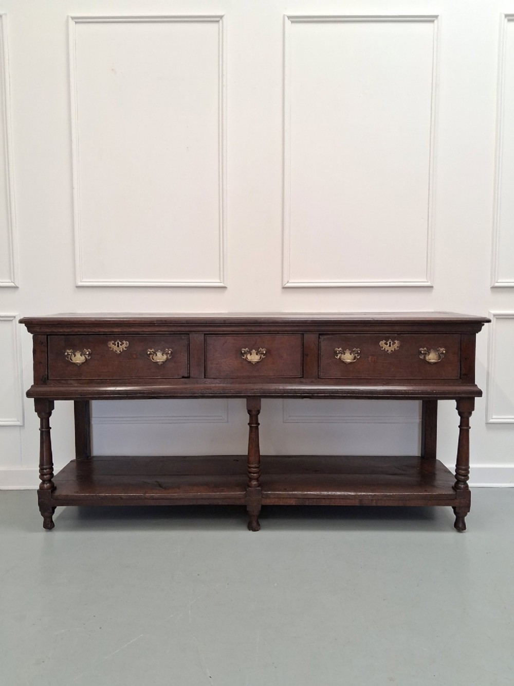 An 18th-century dark oak Georgian dresser base featuring three drawers with brass handles, turned front legs, and a low pot board shelf.