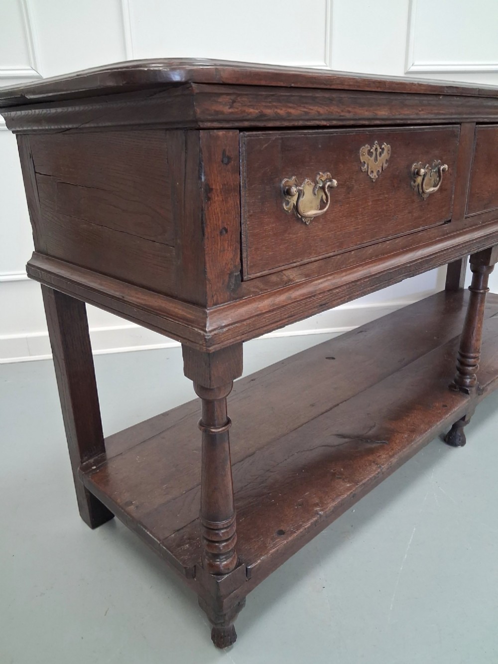 An 18th-century dark oak Georgian dresser base featuring three drawers with brass handles, turned front legs, and a low pot board shelf. View VII