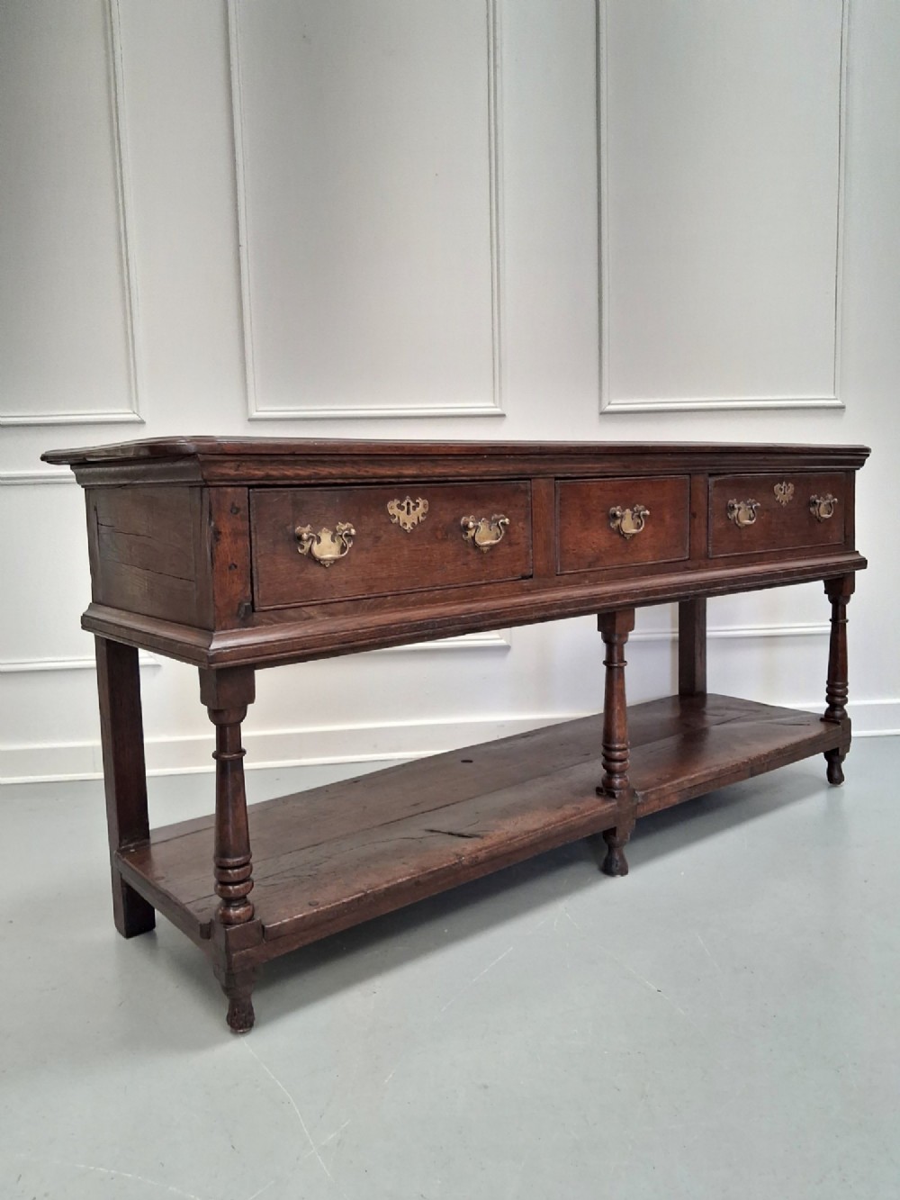 An 18th-century dark oak Georgian dresser base featuring three drawers with brass handles, turned front legs, and a low pot board shelf. View VIII