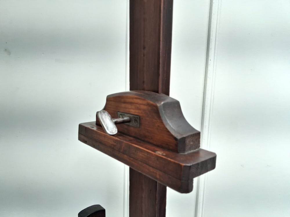 A tall dark pitch pine studio easel featuring an adjustable central mast, lower support shelf, and base castors. View III