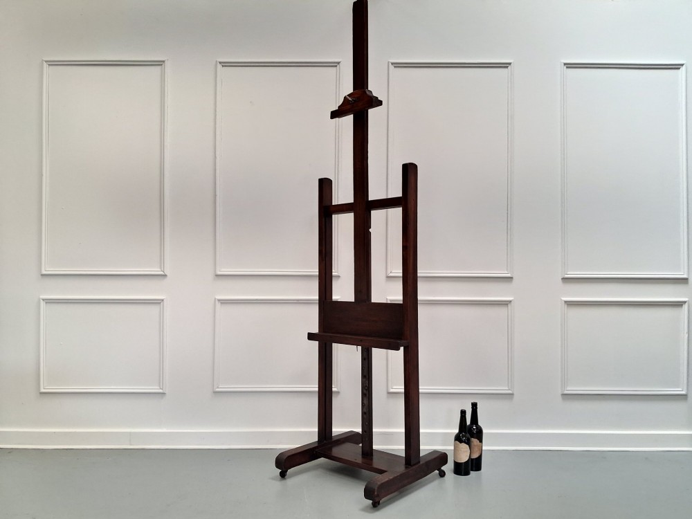 A tall dark pitch pine studio easel featuring an adjustable central mast, lower support shelf, and base castors. View II
