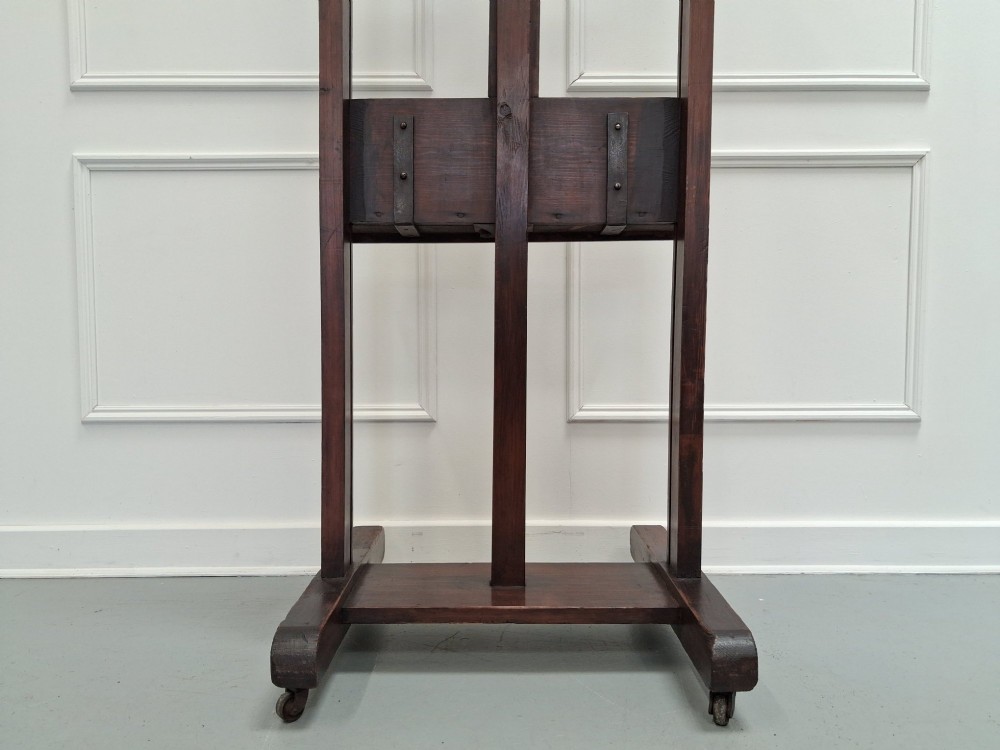 A tall dark pitch pine studio easel featuring an adjustable central mast, lower support shelf, and base castors. View V