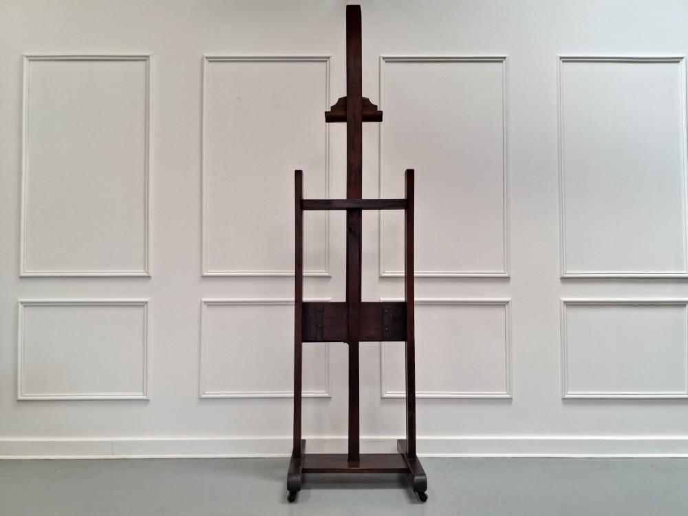 A tall dark pitch pine studio easel featuring an adjustable central mast, lower support shelf, and base castors.