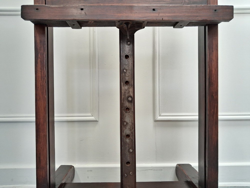 A tall dark pitch pine studio easel featuring an adjustable central mast, lower support shelf, and base castors. View VI