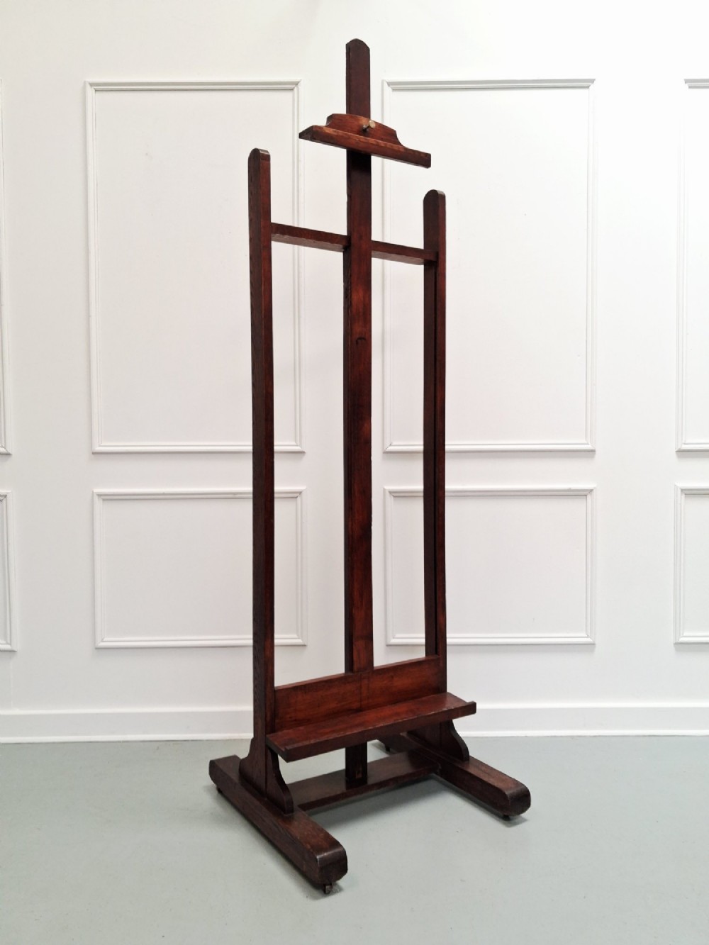 A tall dark oak studio easel featuring an adjustable central mast, upper clamp, solid lower shelf, and a heavy H-frame base.