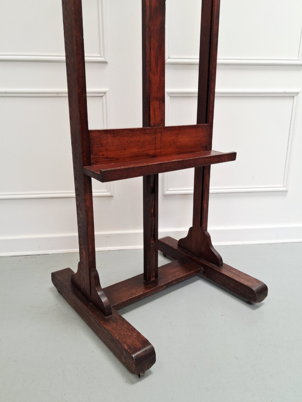 A tall dark oak studio easel featuring an adjustable central mast, upper clamp, solid lower shelf, and a heavy H-frame base. View III