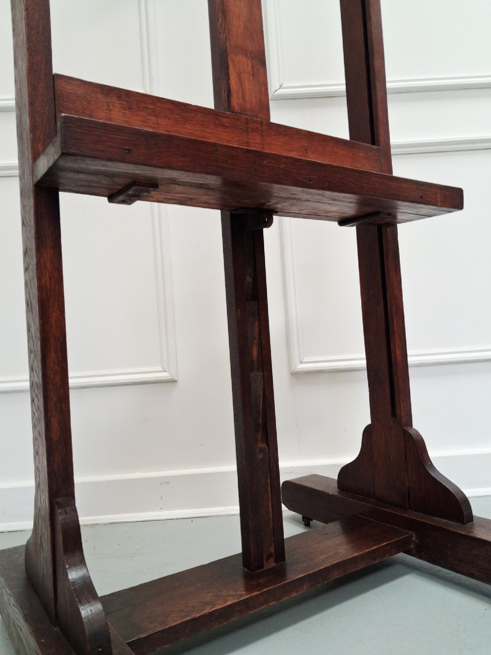 A tall dark oak studio easel featuring an adjustable central mast, upper clamp, solid lower shelf, and a heavy H-frame base. View IV