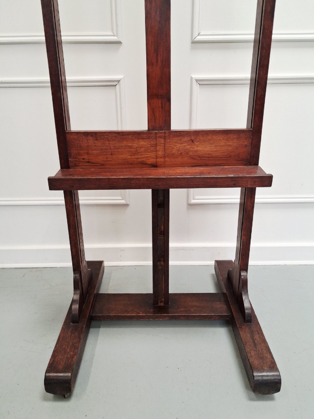 A tall dark oak studio easel featuring an adjustable central mast, upper clamp, solid lower shelf, and a heavy H-frame base. View V