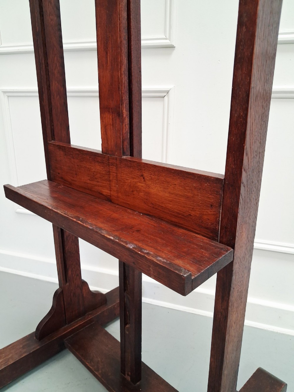 A tall dark oak studio easel featuring an adjustable central mast, upper clamp, solid lower shelf, and a heavy H-frame base. View VI