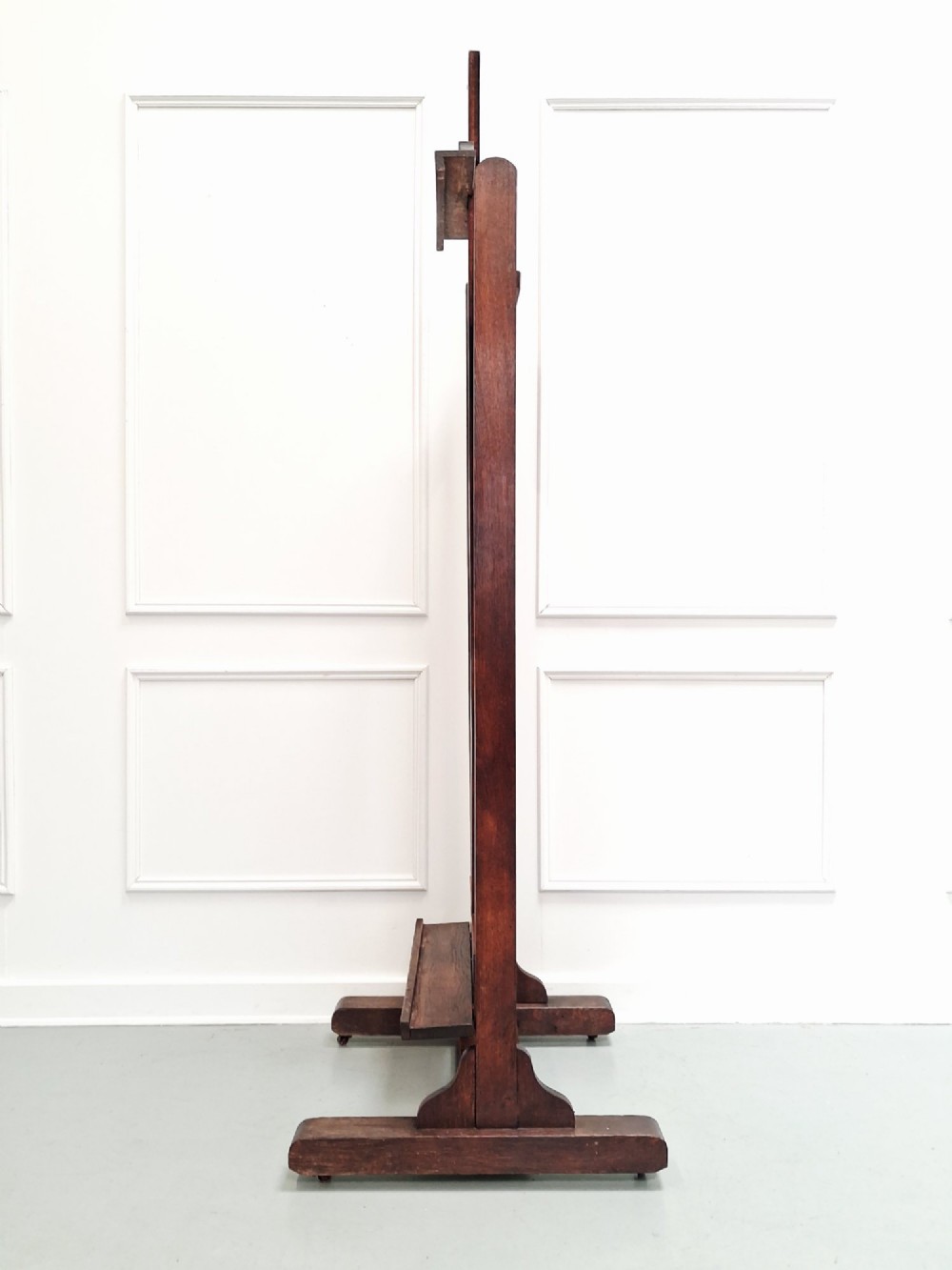 A tall dark oak studio easel featuring an adjustable central mast, upper clamp, solid lower shelf, and a heavy H-frame base. View VII