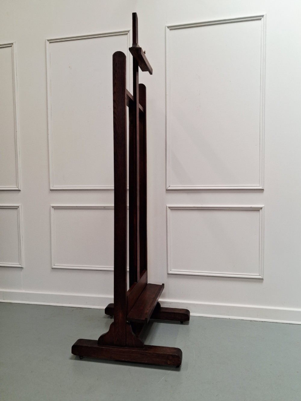 A tall dark oak studio easel featuring an adjustable central mast, upper clamp, solid lower shelf, and a heavy H-frame base. View X