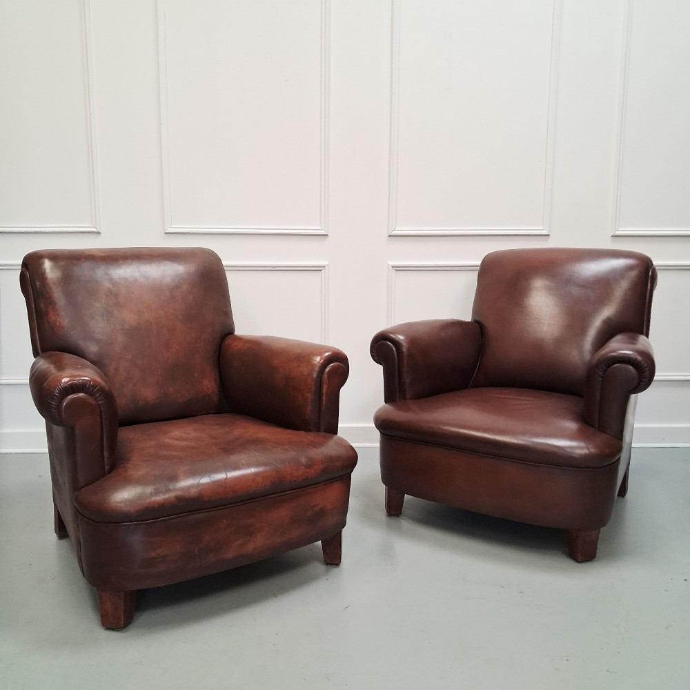A pair of 1930s French club chairs upholstered in warm brown leather with rolled arms and short wooden feet.