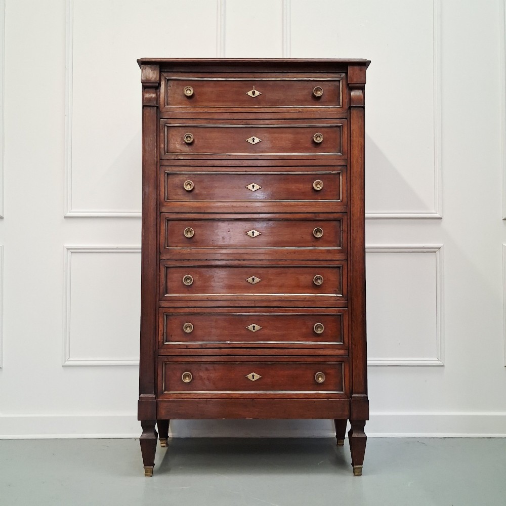 An antique French mahogany semainier tallboy featuring seven drawers with round brass pulls and diamond-shaped brass escutcheons set on short turned feet.