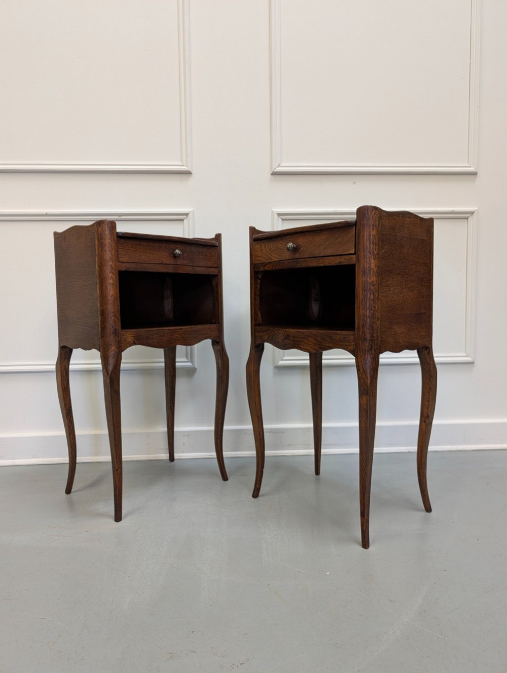 A pair of French dark oak bedside tables featuring slender cabriole legs, an open shelf alcove, and a single drawer with a metal knob.