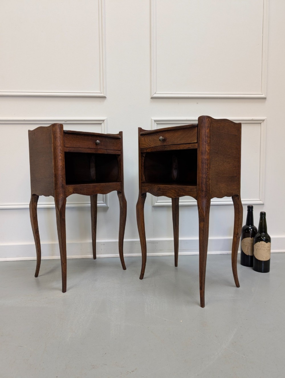 A pair of French dark oak bedside tables featuring slender cabriole legs, an open shelf alcove, and a single drawer with a metal knob. View VI