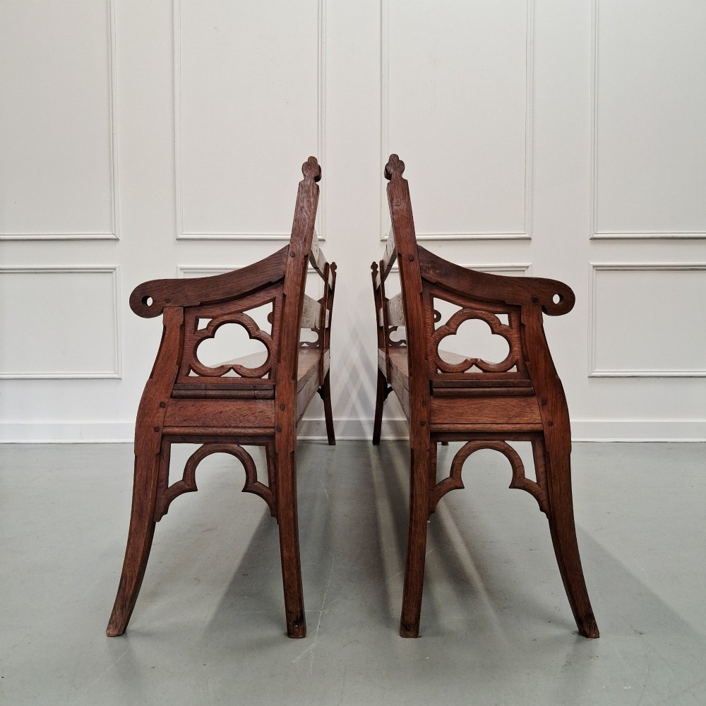 Profile view of a pair of antique Gothic Revival solid oak gallery benches featuring carved trefoil motifs and curved armrests.