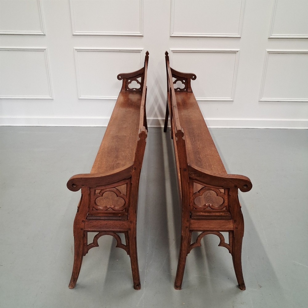 Profile view of a pair of antique Gothic Revival solid oak gallery benches featuring carved trefoil motifs and curved armrests. View II